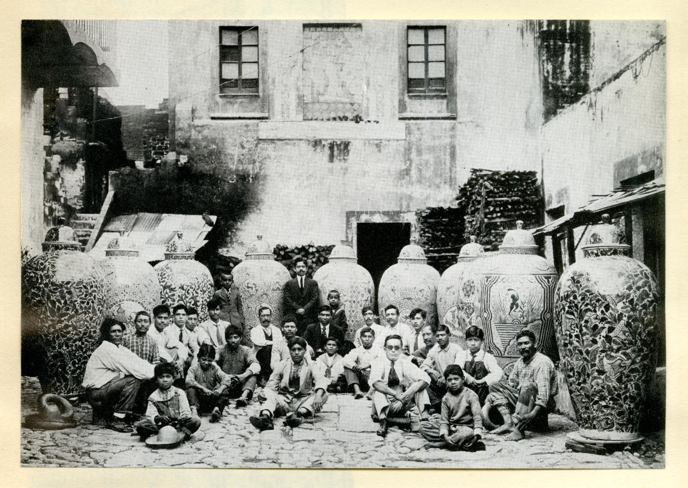 27 men and boys gather in front of 9 oversize vessels taller than the standing man.