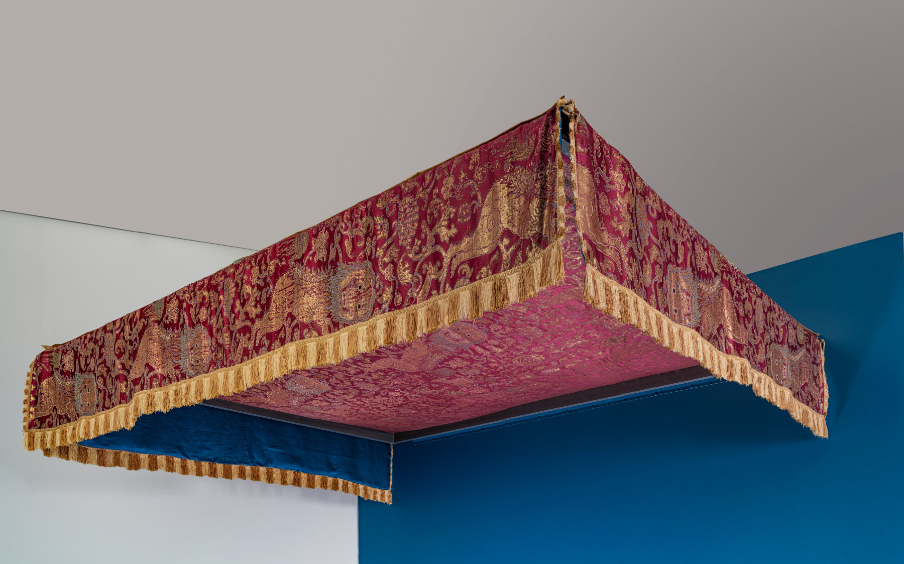 A rectangle juts out from a blue wall. The structure is covered with red cloth embellished with gold fringe and animal and floral motifs.