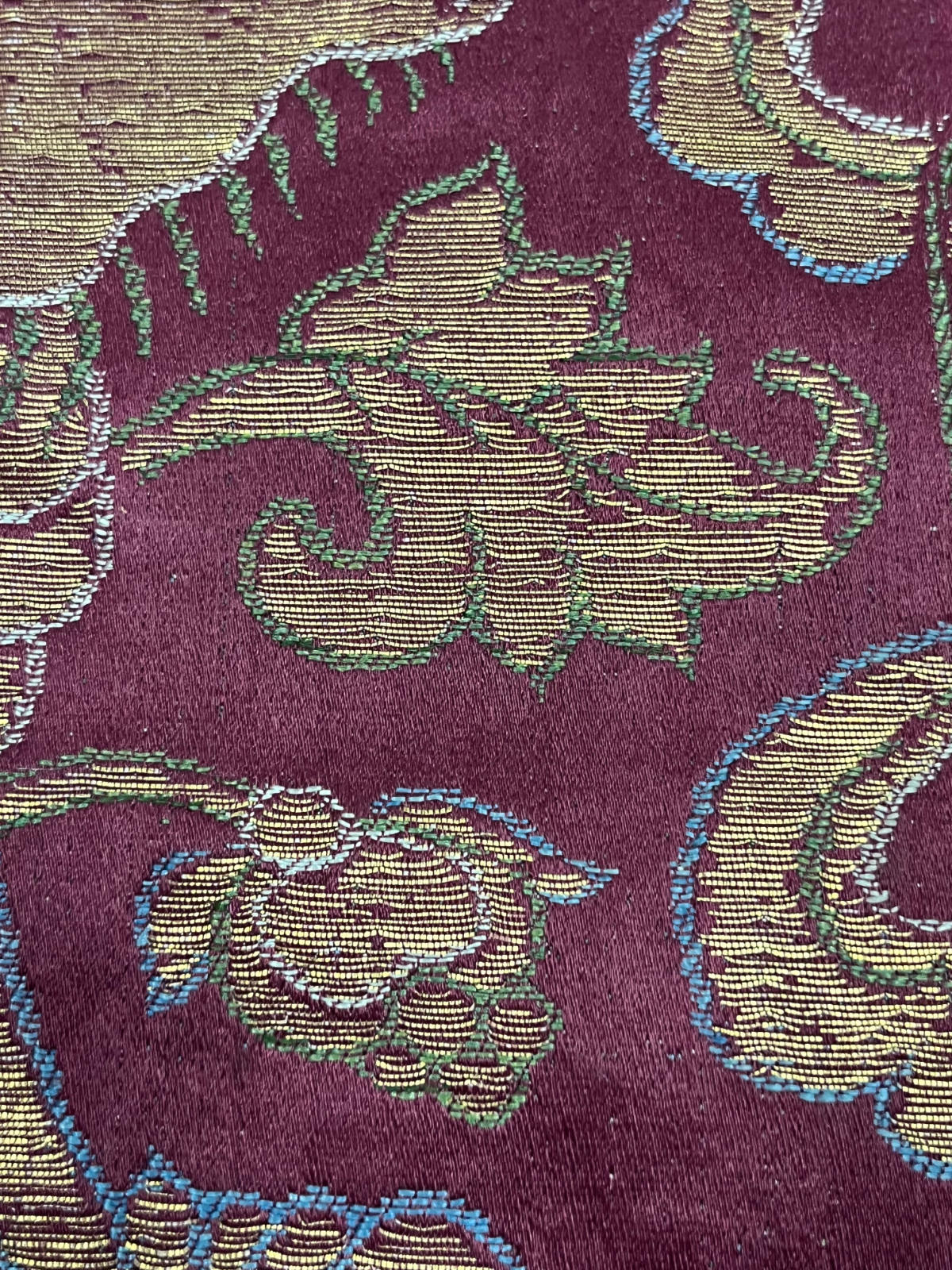 A close-up view of floral motifs on a textile. The cloth is burgundy, the leaves and flowers have green and blue edges and are filled in with yellow thread.