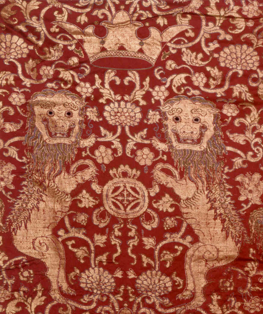 A close-up view of a red textile adorned with gold lions, a crown at center, and floral motifs throughout.