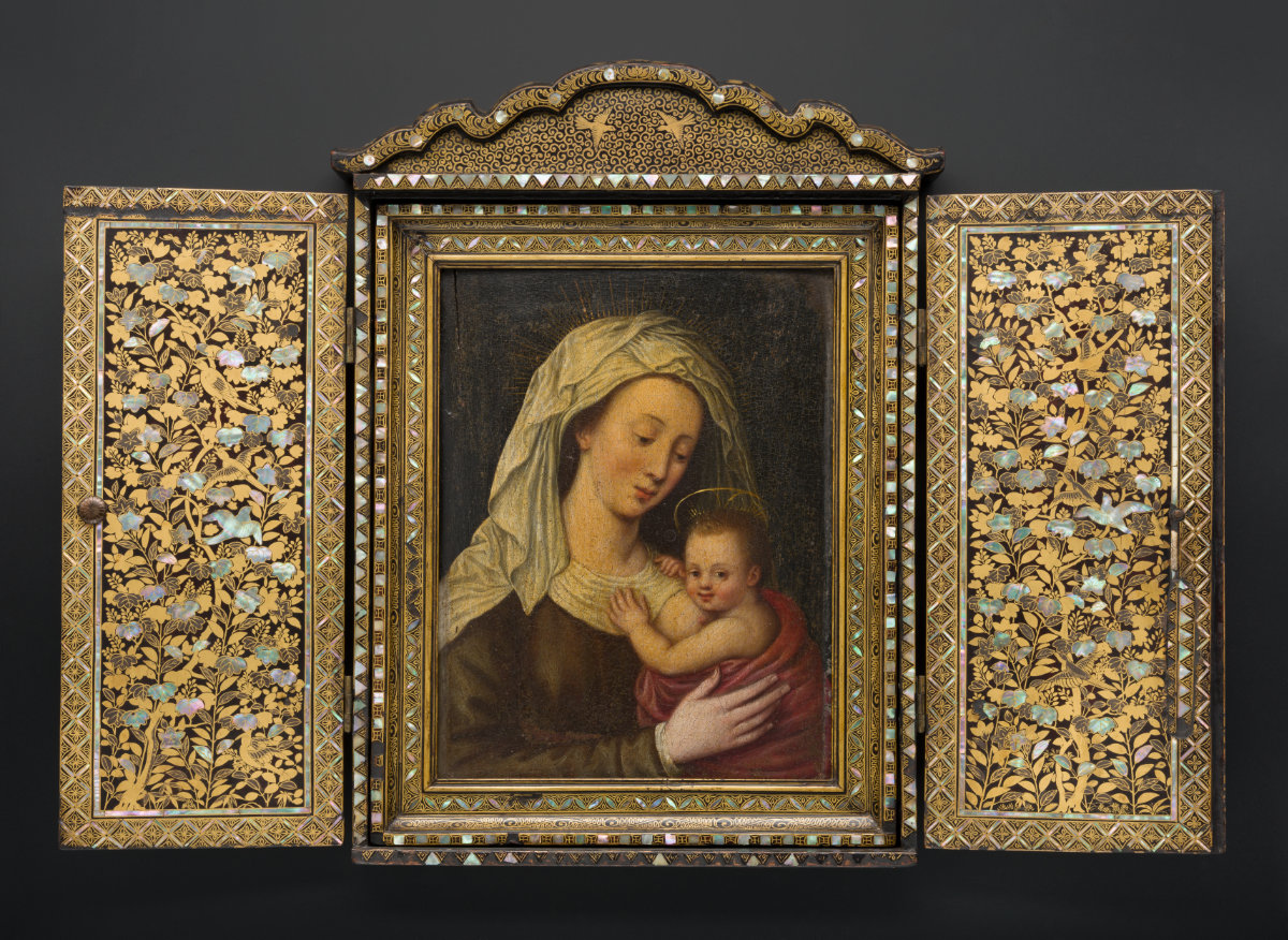 A painting of a woman with a white head covering and gold halo holds an infant swaddled in red cloth and a halo. The painting is inside a gold shrine with floral motifs and two birds at top.