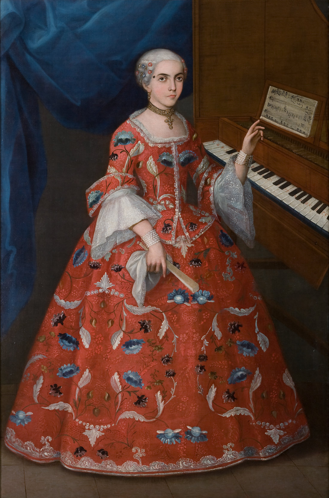 A woman with white hair in a floor-length red dress points toward the harpsichord next to which she stands.