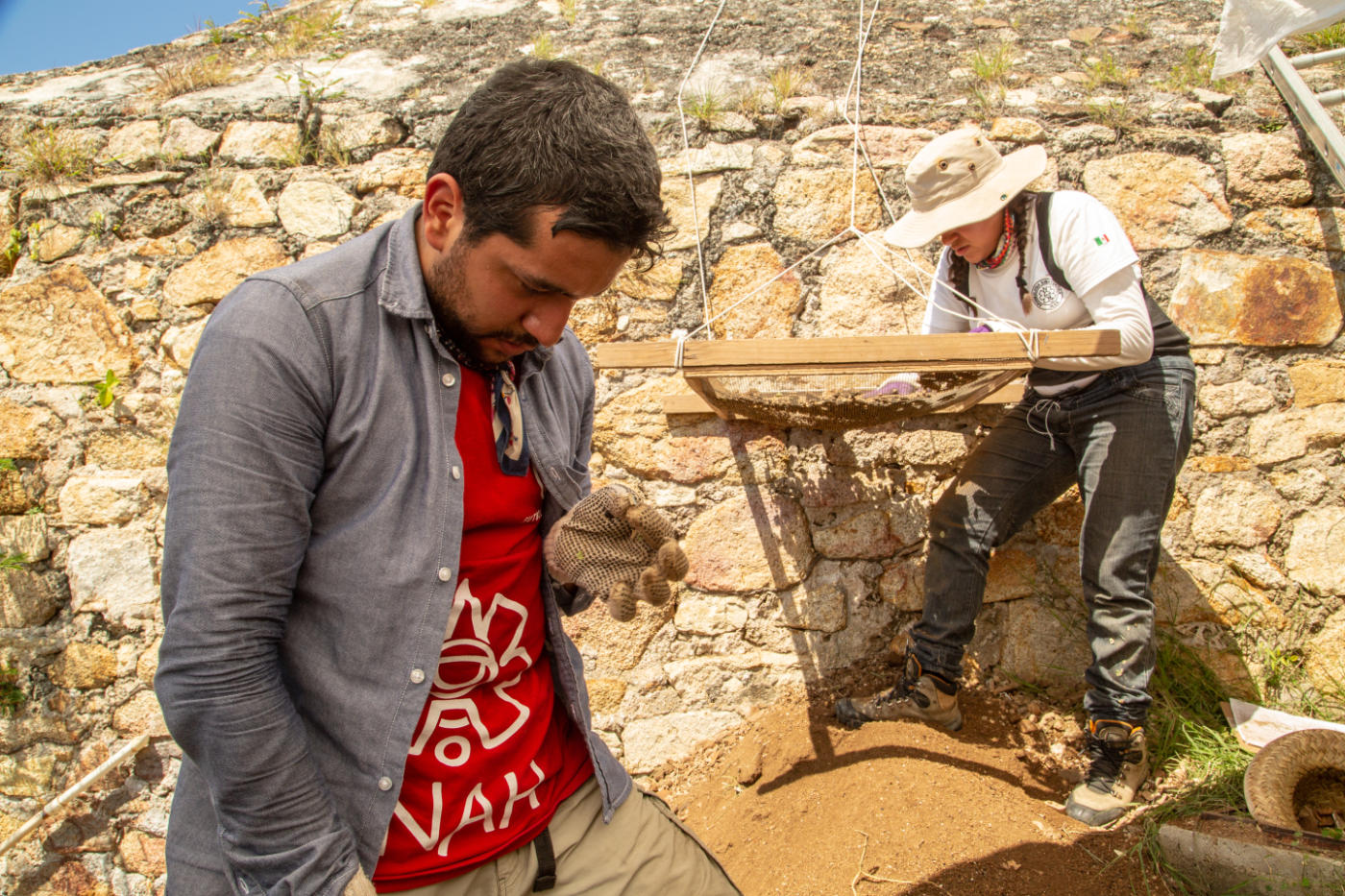 Two people are in front of a stone wall, one looking down at their hand and one bending over a wooden structure suspended from ropes.