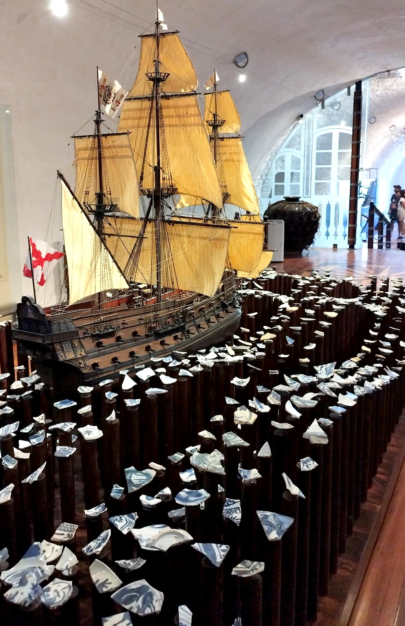 A model galleon with sails unfurled in a museum gallery surrounded by blue-and-white shards.