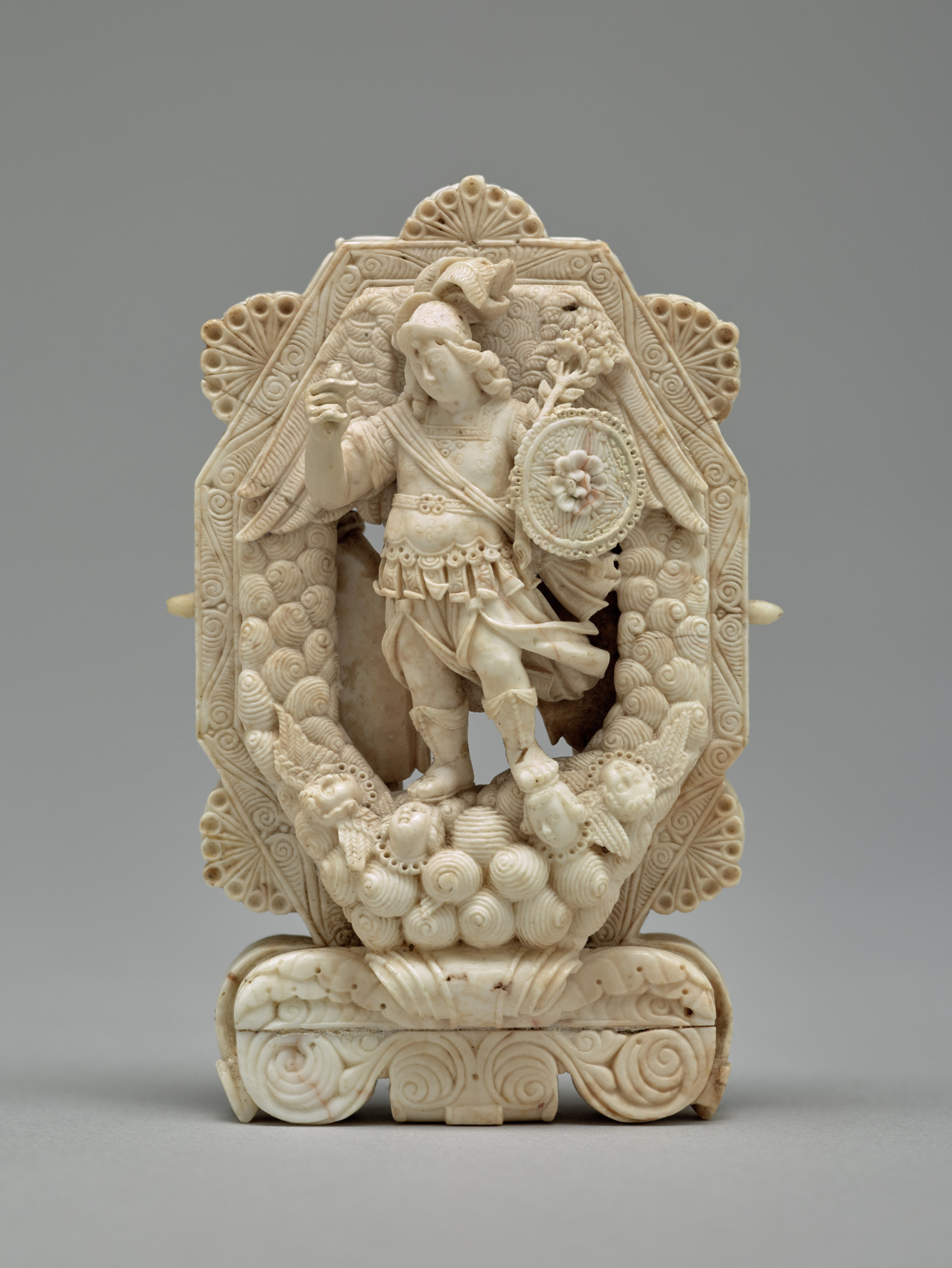 A white sculpture featuring a figure holding a shield standing on two disembodied heads and surrounded by geometric and swirl motifs.