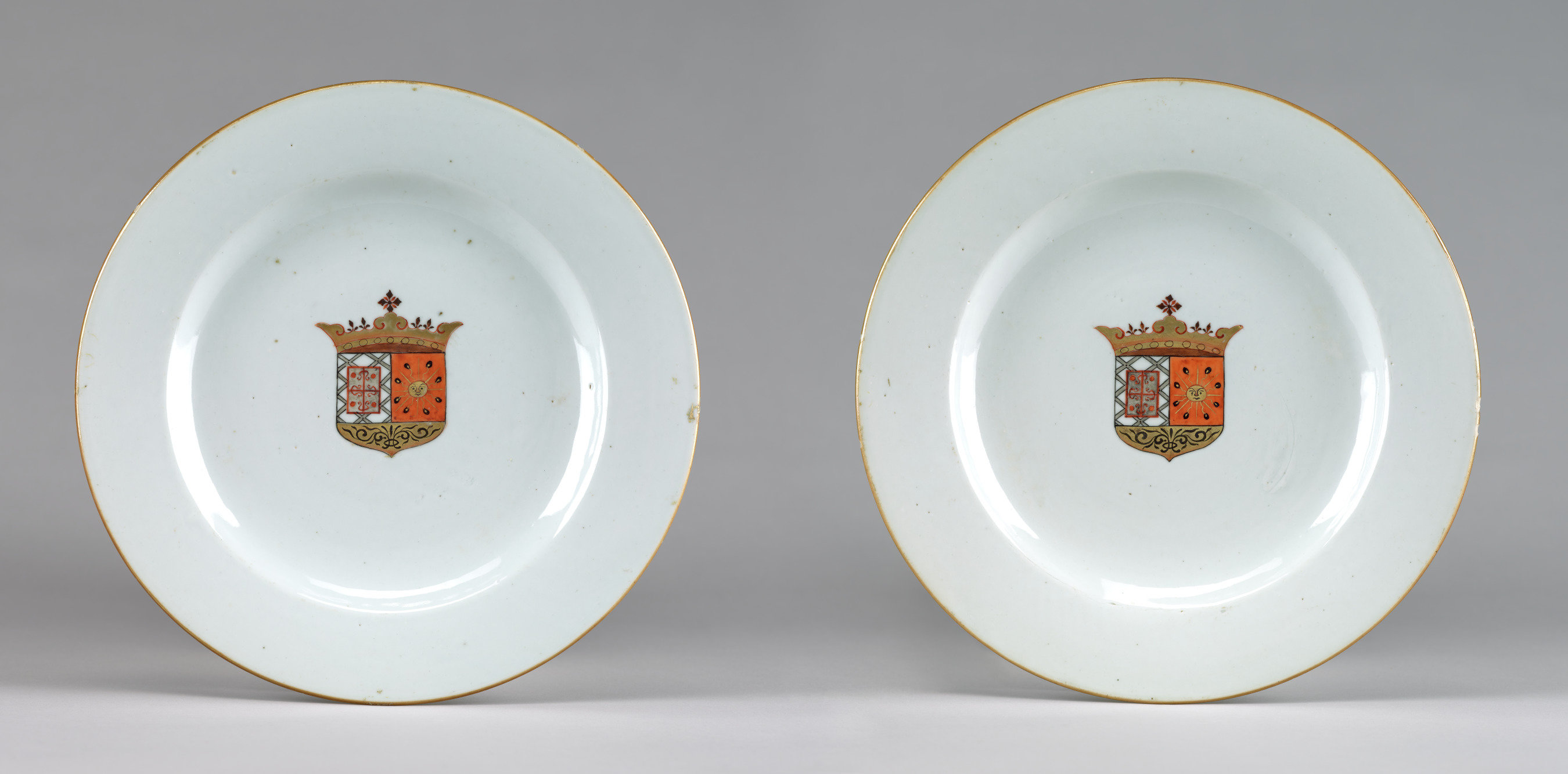 Two white plates with gold borders each feature a coat of arms at center comprising crown, cross, and sun motifs.