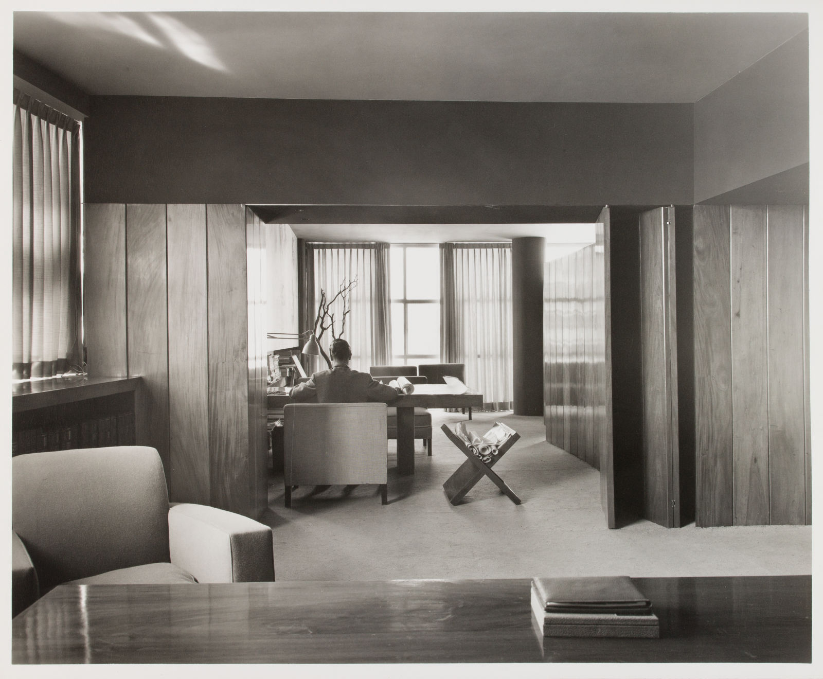 A black and white photograph of Juan Sordo Madaleno working in his studio. Madaleno sits in a different room from us, framed by the wood paneled doorway and with his turned towards us.