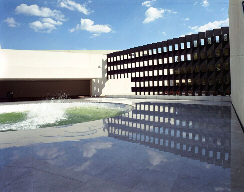 A photograph of a wide exterior space with an in ground water fountain and modernist black gateway with squared holes and  an open space for passersby to the left.